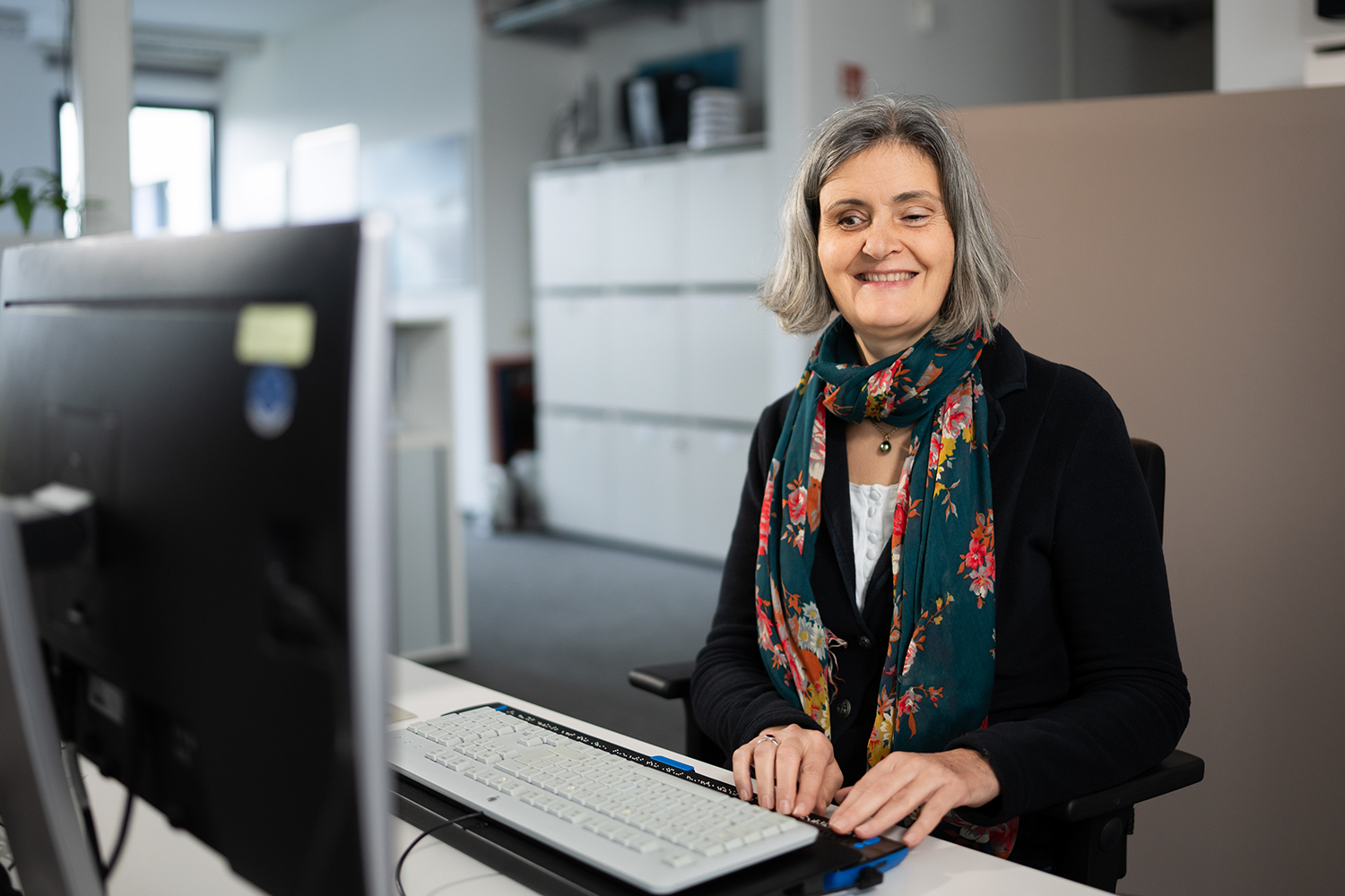 Eine blinde Kollegin bei der Arbeit am Computer mit technischem Hilfsmittel