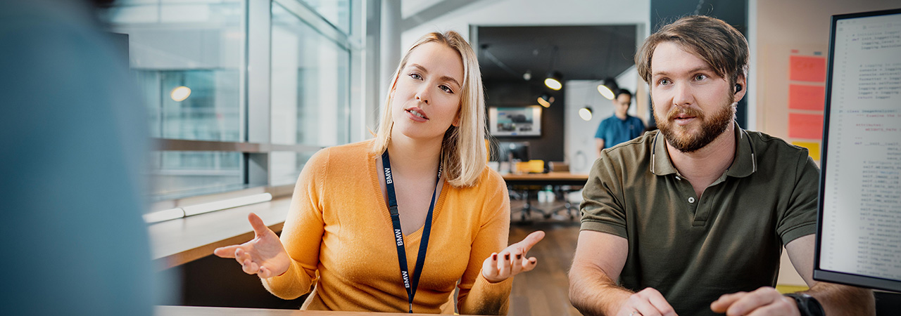 Three colleagues at the BMW Group are having a collaborative conversation in a modern office setting.