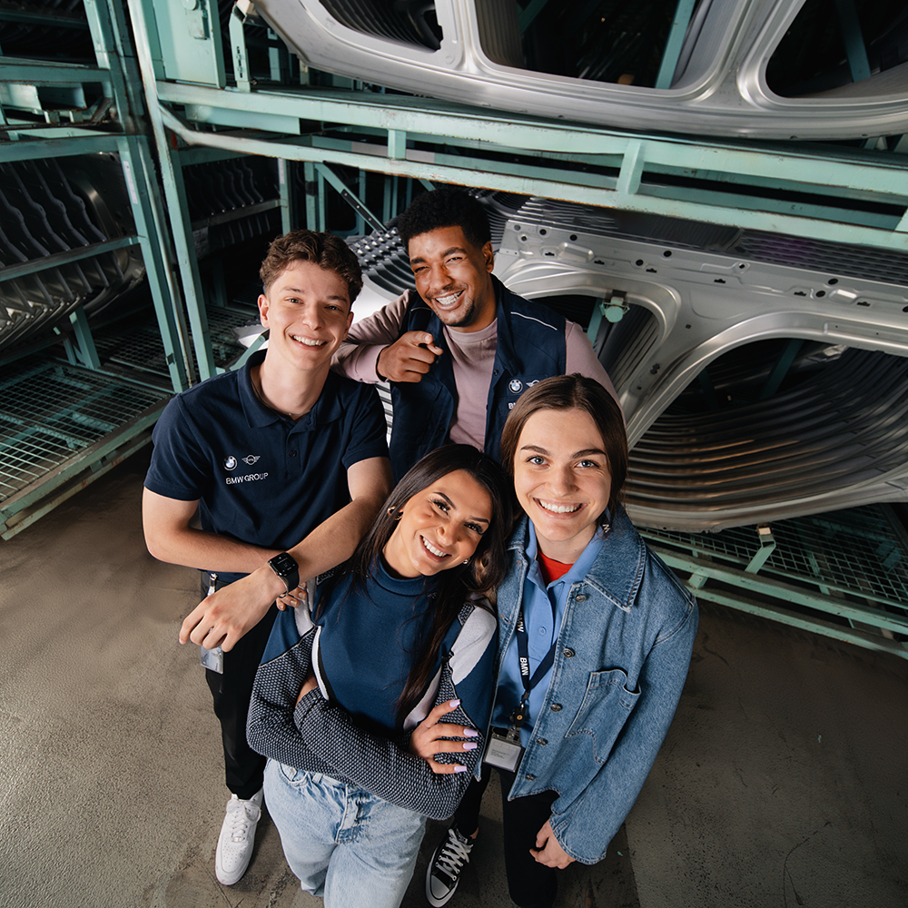 A diverse group of young professionals smiling together in a manufacturing environment.