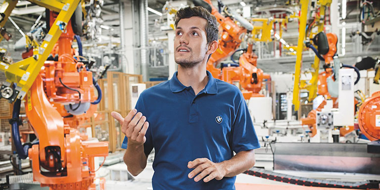 The picture shows a young man, working in Engineering and R&D at BMW, explaining something in a car manufacturing hall.
