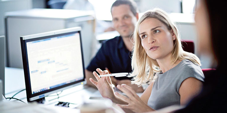 The picture shows a woman working in IT at BMW explaining something in front of a computer.